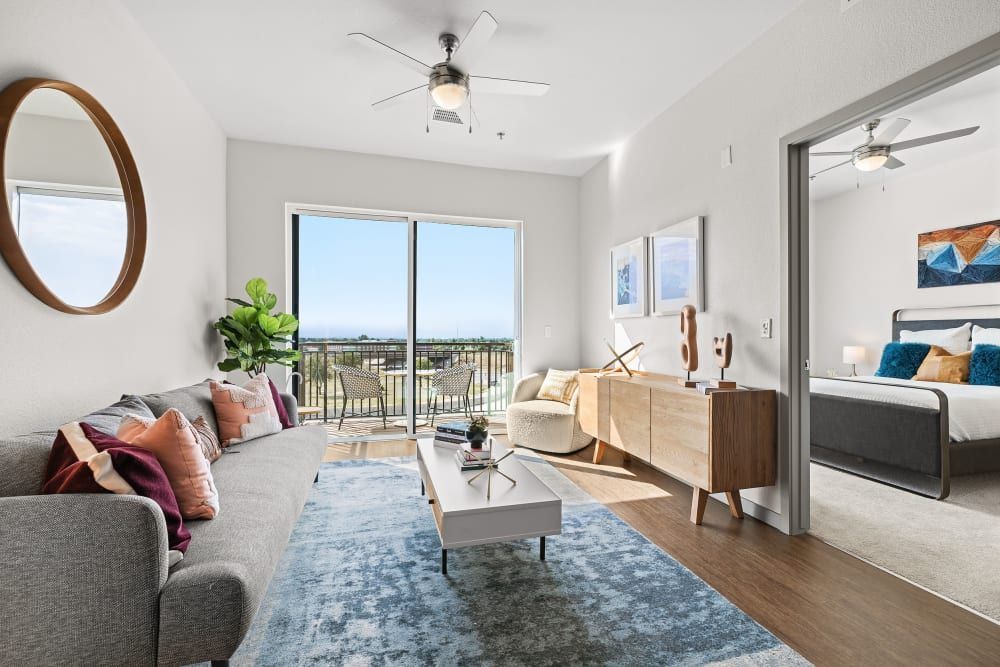 Modern and bright one-bedroom apartment interior with open floor plan. Features a living area with a gray couch, multicolored throw pillows, and a round mirror on the wall, leading to a balcony with outdoor seating. A bedroom is visible in the background with a bed adorned with blue and beige bedding and a wooden nightstand. The decor includes indoor plants, wall art, and a stylish mid-century modern sideboard with decorative items. Large windows provide abundant natural light and a view of the exterior at Marq Iliff Station in Aurora, CO.