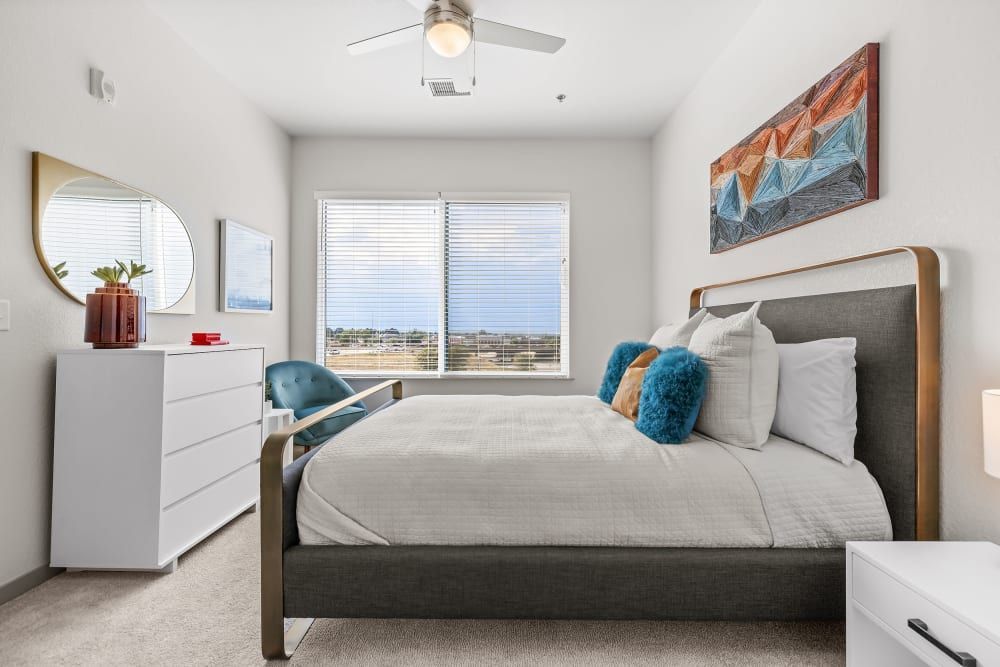 A modern, well-lit bedroom featuring a large bed with a gray upholstered headboard and white bedding, accented by blue pillows. A white dresser with a circular mirror above it is on the left, and a blue armchair sits near the bed. Abstract artwork hangs on the wall, and the room has a neutral color palette with white walls and a light carpet. A window with horizontal blinds offers a view of the outdoors, inviting natural light into the space at Marq Iliff Station in Aurora, CO.