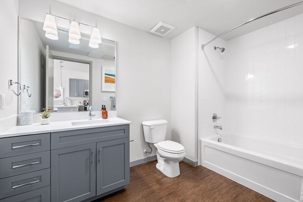 Modern bathroom interior with white walls featuring a large mirror above a gray vanity with a white countertop, undermount sink, and pendant lights. To the right is a white toilet next to a built-in bathtub with a white tile surround and shower. The floor is covered with a patterned brown vinyl at Marq Iliff Station in Aurora, CO.