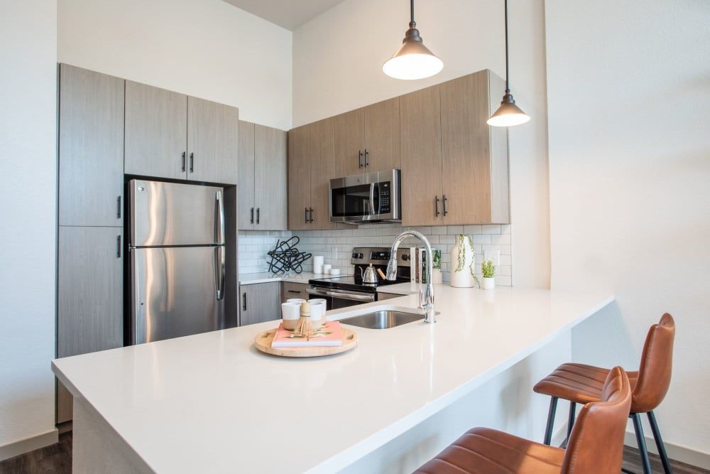 Modern kitchen interior with gray cabinets, stainless steel refrigerator, black stove and microwave, and a white countertop. Two pendant lights hang above a breakfast bar where two brown leather bar stools are placed. Decorative plants and a serving tray with coffee cups add a cozy touch to the space at Marq Iliff Station in Aurora, CO.