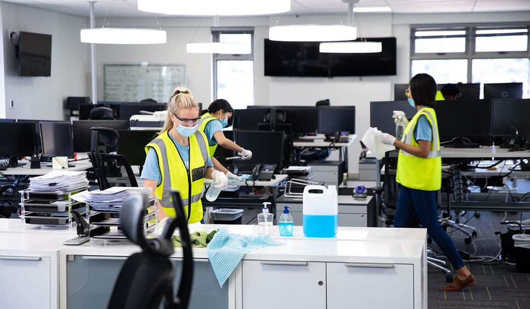 Workers in safety vests cleaning a modern office with desks, computers, and supplies.