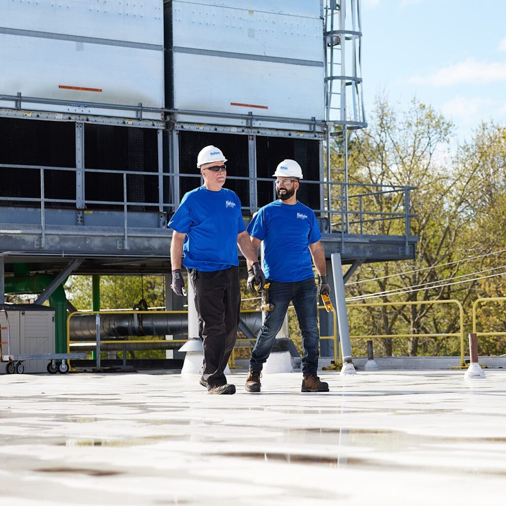 Two workers in blue shirts and hard hats walk on a rooftop near large industrial equipment.
