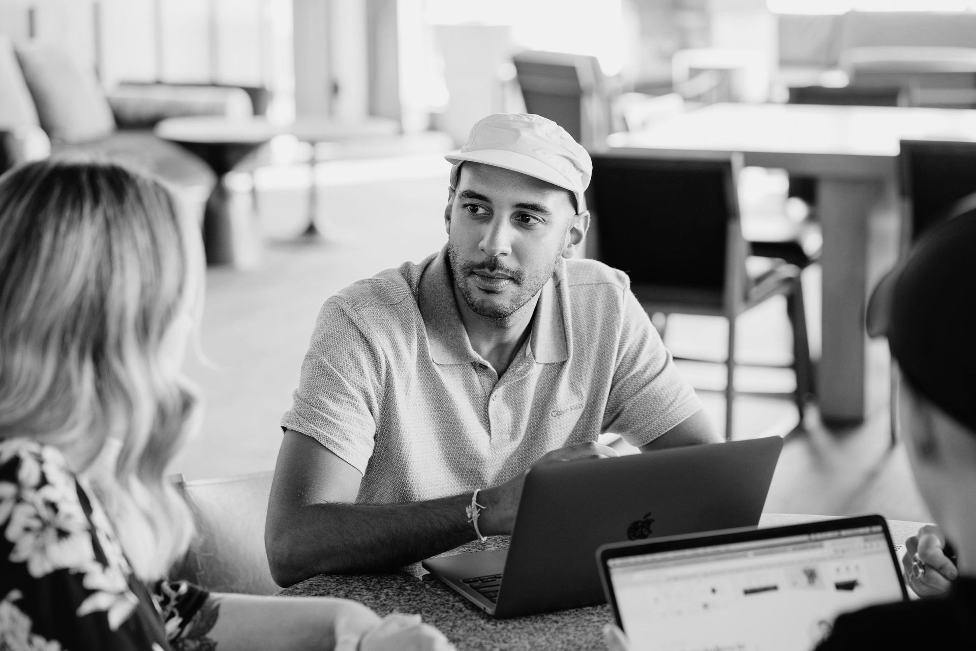 Man in a cap, working on a laptop with two others at a table in a bright, modern setting.
