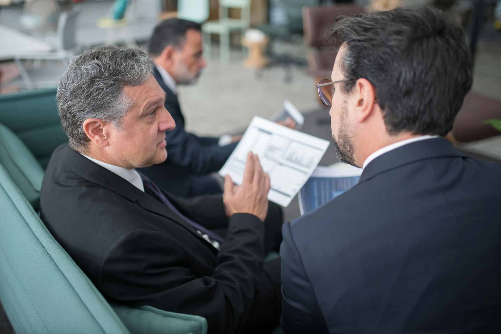 Three men in suits reviewing documents on a teal couch; one points while speaking.