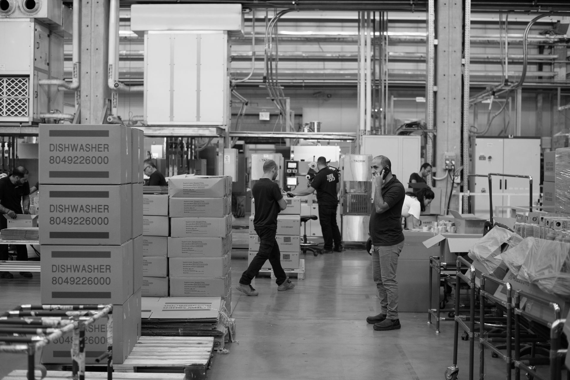 Warehouse interior with workers. Man on phone, boxes stacked, other people loading.