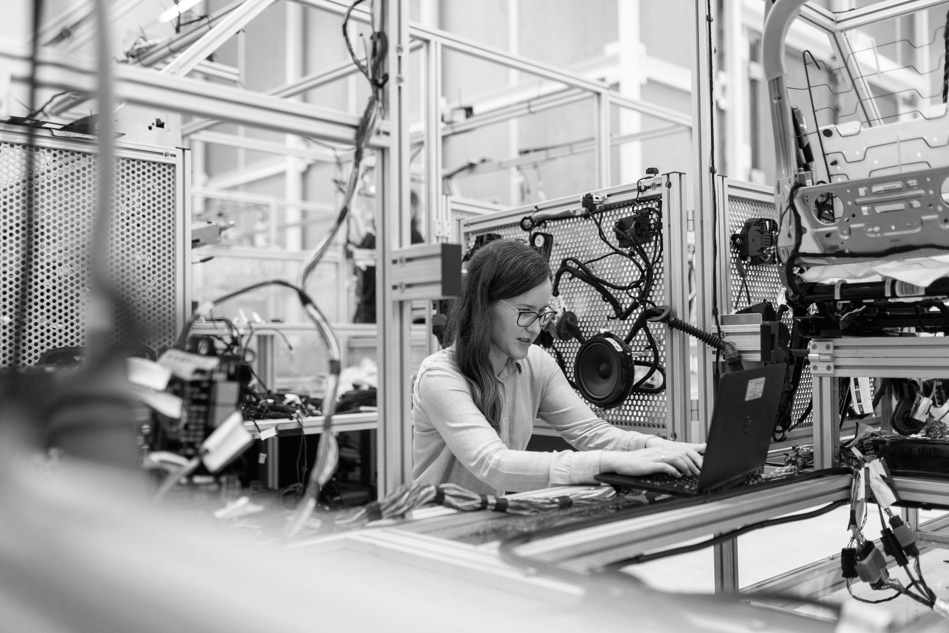Woman working on a laptop in an industrial lab setting, surrounded by machinery and wires.