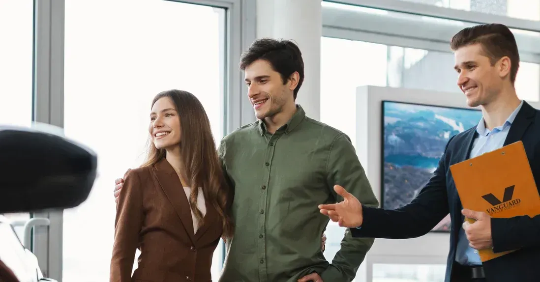 Couple smiles while looking at a car with a salesperson. They are in a car showroom.