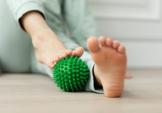 Persona que utiliza una pelota de masaje verde con púas en el pie mientras está sentada en un piso de madera.
