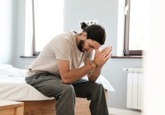 Hombre con la cabeza inclinada y las manos juntas en oración, sentado en una cama en un dormitorio, mostrando una expresión pensativa.