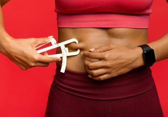 Mujer midiendo su grasa corporal con un calibrador. Fondo rojo, con sujetador deportivo y reloj inteligente.