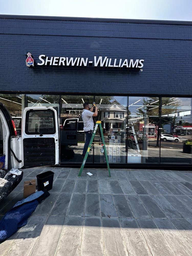 A person on a ladder is installing a sign for Sherwin-Williams on a storefront.