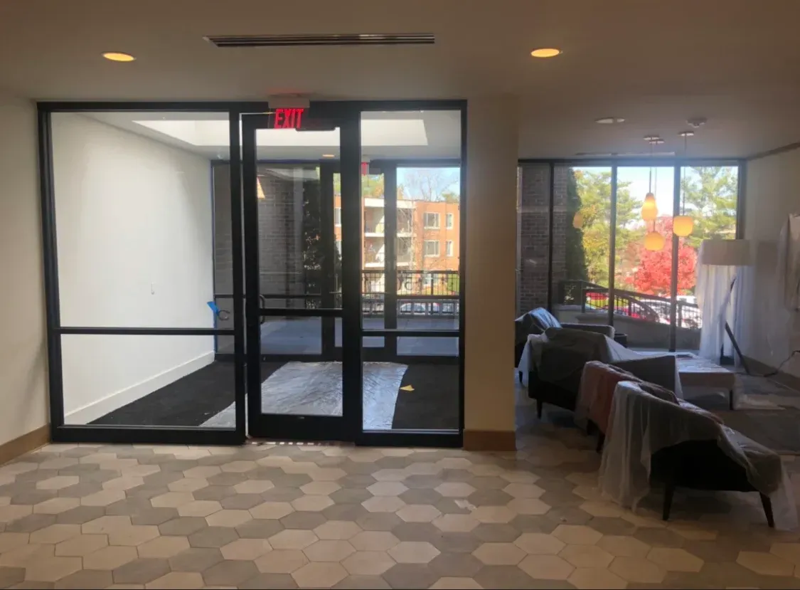 Interior view of lobby with black-framed glass doors and windows; chairs covered in plastic; patterned floor.