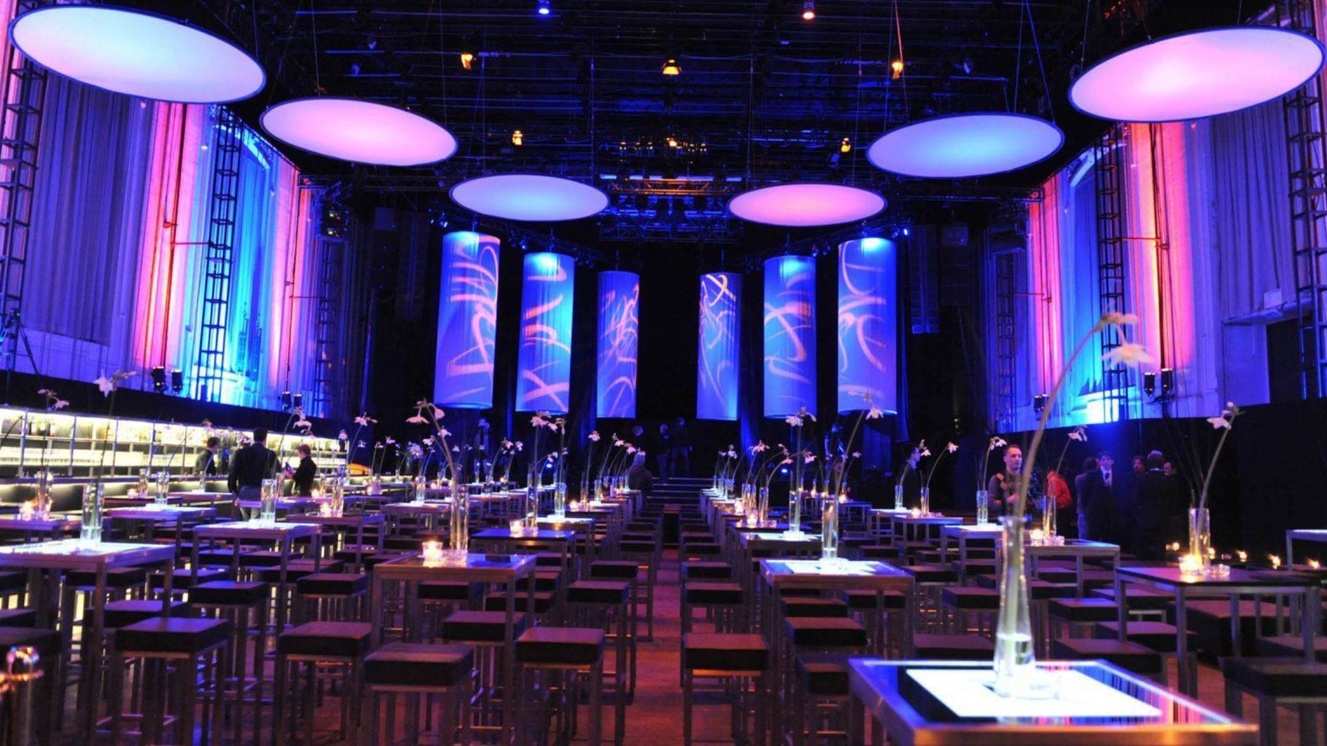 Elegant banquet hall with purple-blue lighting, round tables, chairs, and tall illuminated columns