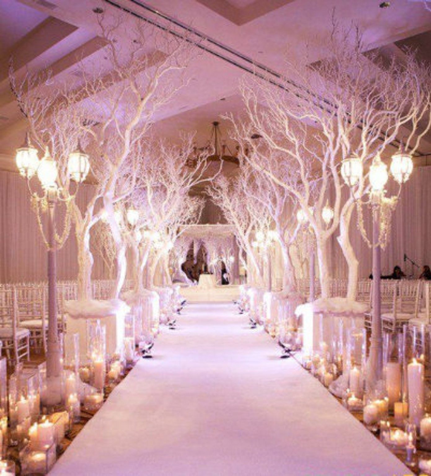 Elegant wedding aisle lined with white trees, candles, and warm lights in a softly lit venue