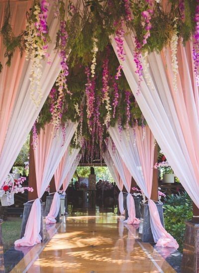 Elegant wedding aisle with white drapes, pink flowers, and hanging greenery over a reflective floor