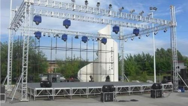 Outdoor concert stage with truss rigging, blue lights, and speakers set up under a clear sky