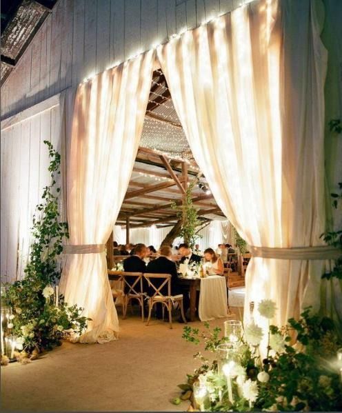 Warm-lit wedding reception through cream drapes, with guests seated at tables in a rustic venue.