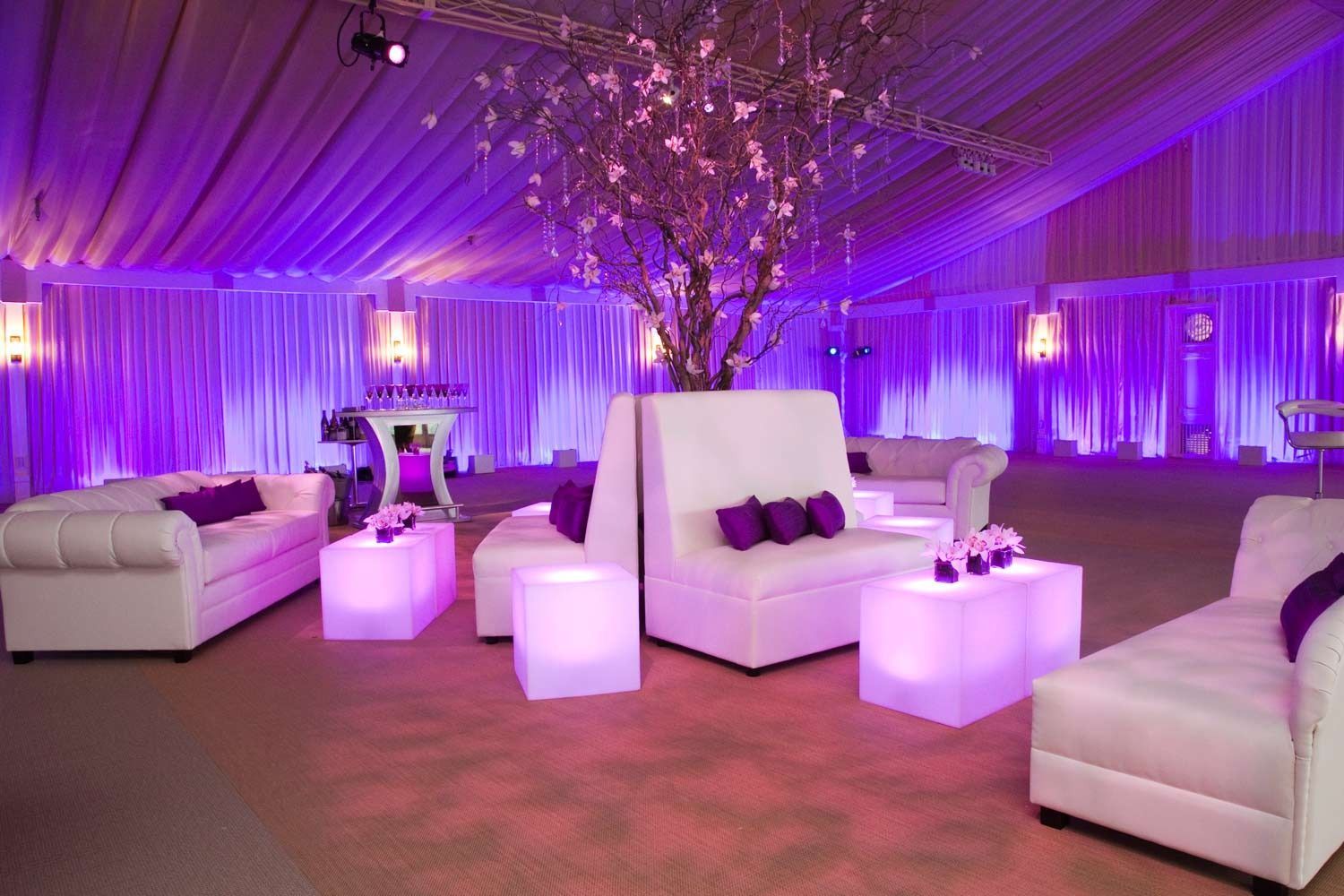 Purple-lit lounge with white sofas and glowing cube tables at an elegant event venue