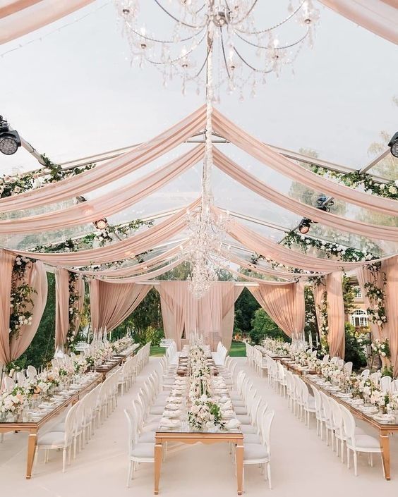 Elegant outdoor wedding reception tent with blush drapes, chandeliers, greenery, and long banquet tables