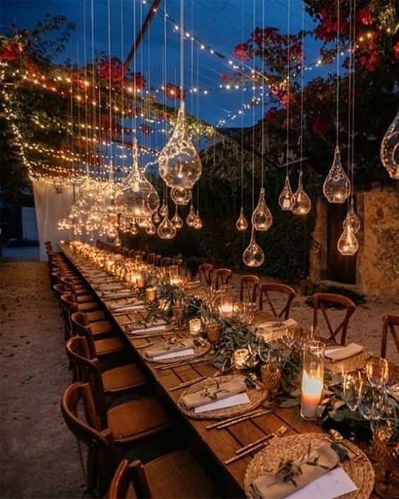 Elegant outdoor dinner table under hanging glass lights at dusk, with warm candlelit decor and greenery.