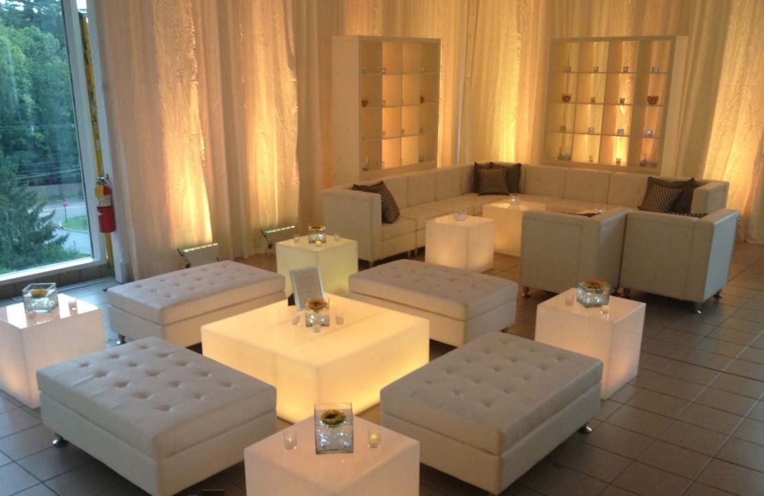 Modern lounge with cream sofas and ottomans, glowing cube tables, and tall curtained windows at dusk