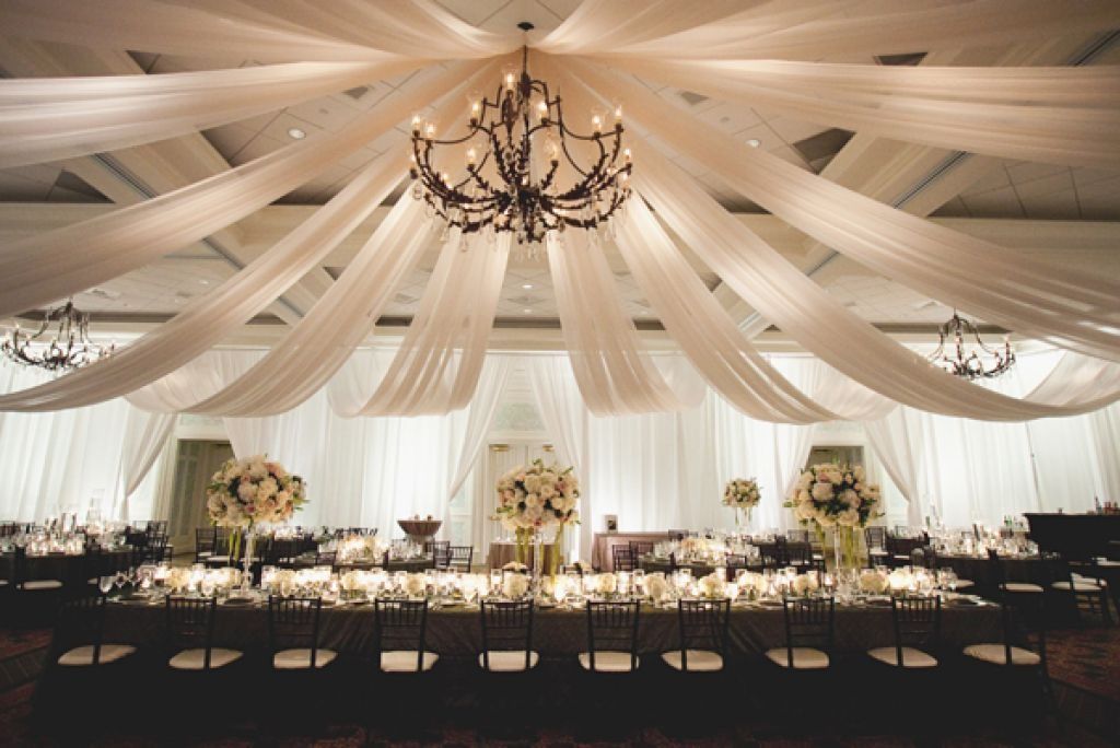 Elegant wedding reception under cream draped ceiling with chandeliers, long floral tables, and black chairs