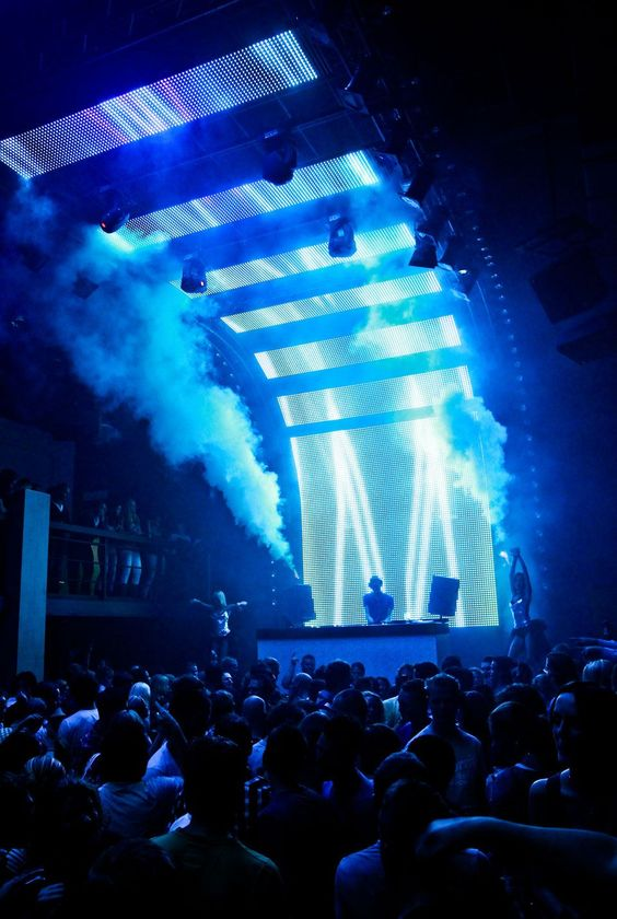 DJ performing on a blue-lit stage with smoke and a cheering crowd in front
