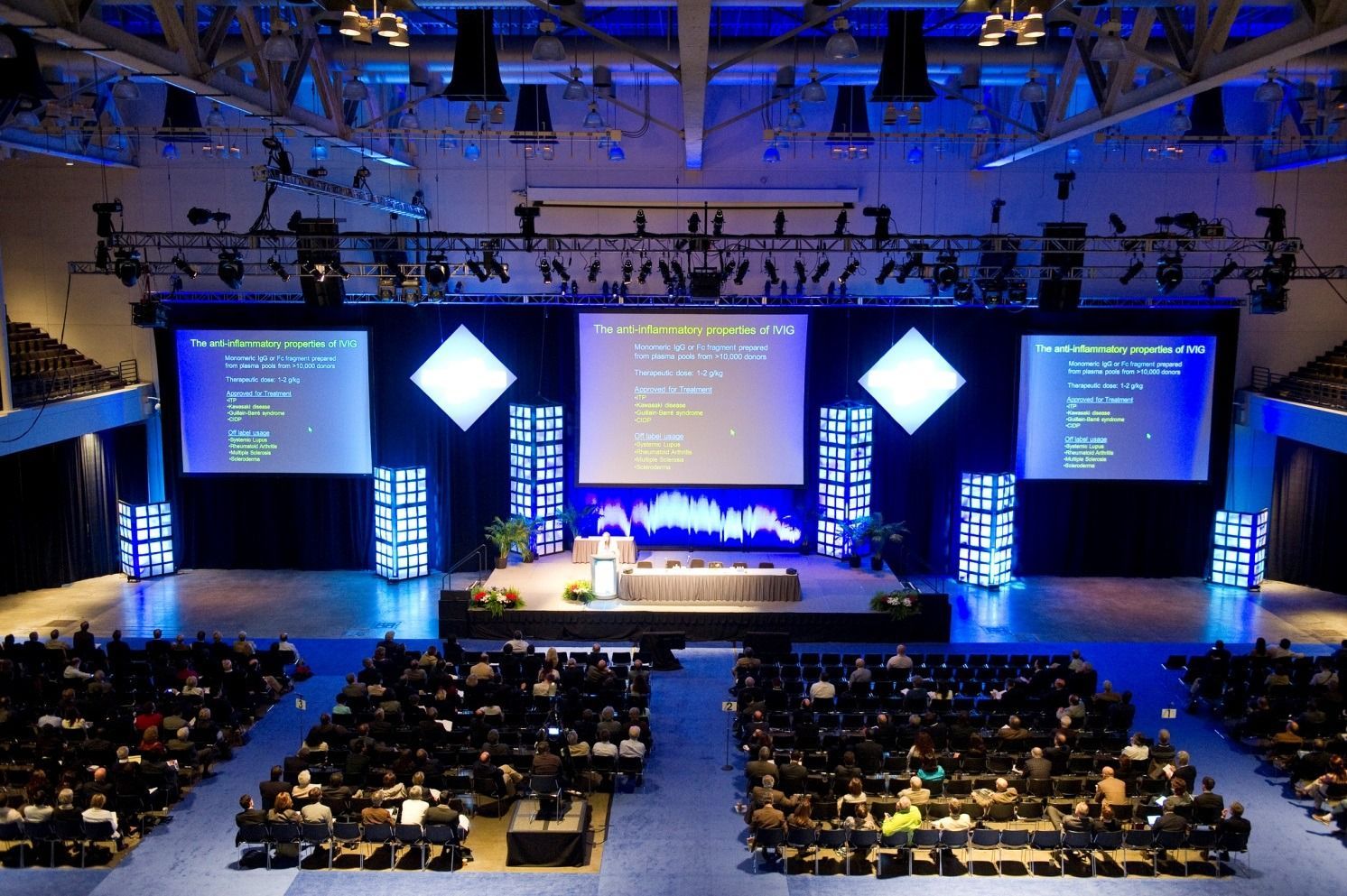 Wide auditorium with a lit stage, large presentation screens, and seated audience at a conference