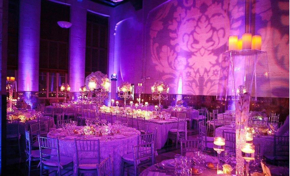 Purple-lit banquet hall with round tables, chairs, candles, and ornate wall projections