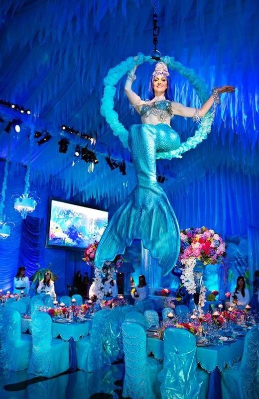 Blue mermaid performer on a stage, surrounded by matching blue decor and a feathered hoop backdrop