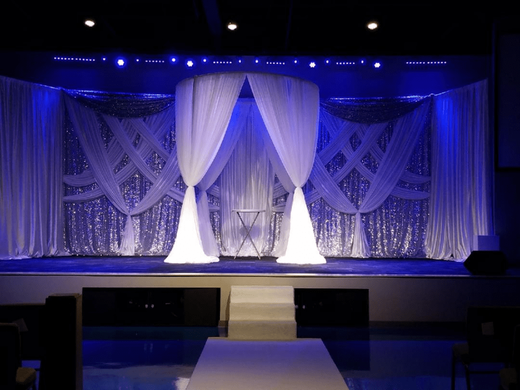 Blue-lit stage with white draped curtains and a centered staircase, set for a performance