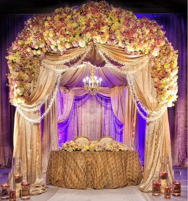 Elegant floral canopy with gold drapes, purple lighting, and a round table centerpiece at a formal event stage