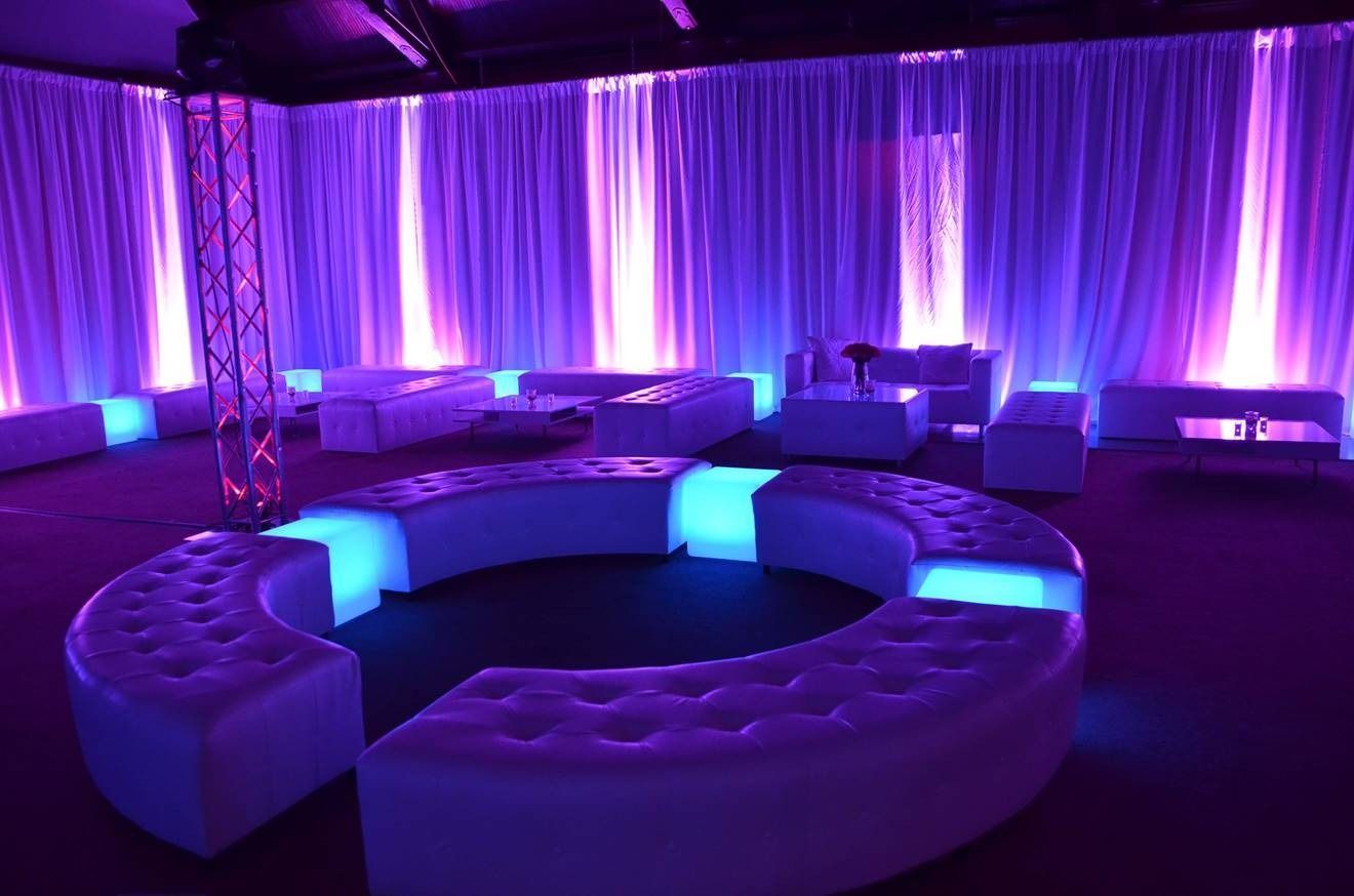Purple-lit lounge with curved white seating and glowing blue tables before draped curtains