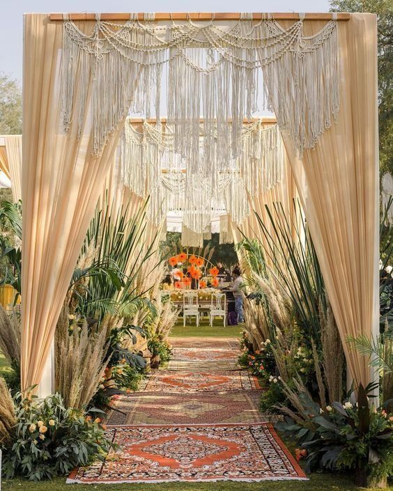 Elegant outdoor wedding aisle with draped beige curtains, lush greenery, and patterned rugs leading to a floral altar