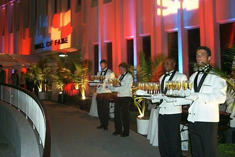 Servers in white jackets holding trays at a red-lit event entrance