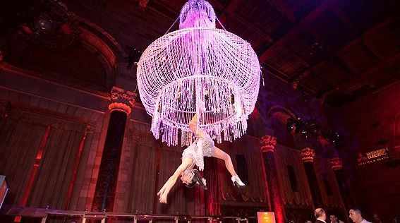 Aerial performer hanging beneath a glowing crystal chandelier in a grand red-lit hall