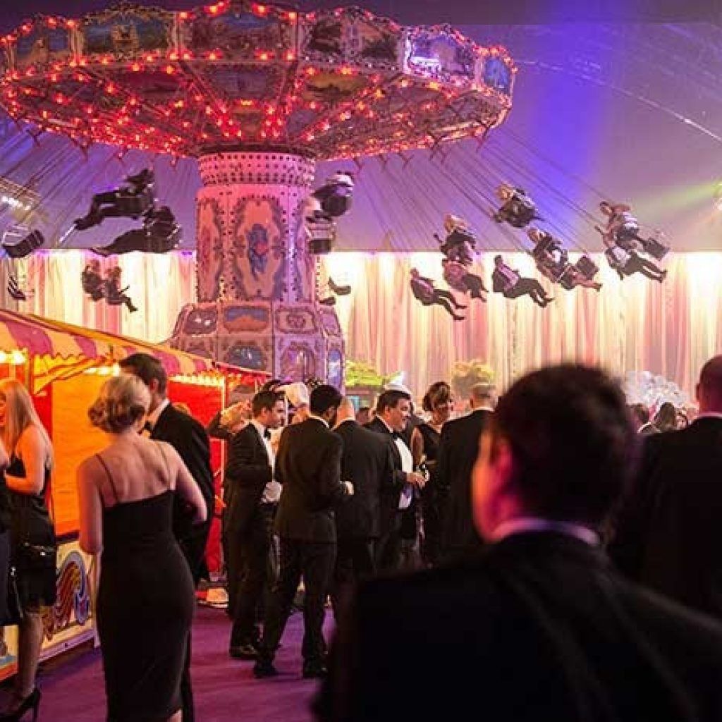 Guests in formalwear at a brightly lit carnival swing ride under a colorful tent at night