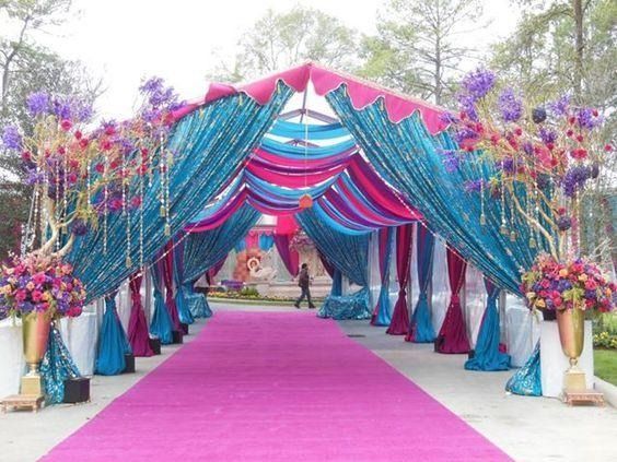 Decorative event walkway with pink carpet and colorful draped canopy, lined with floral arrangements