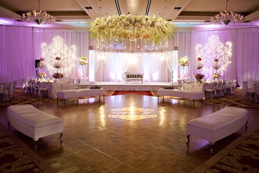 Elegant indoor event venue with white drapes, purple lighting, and floral center stage.