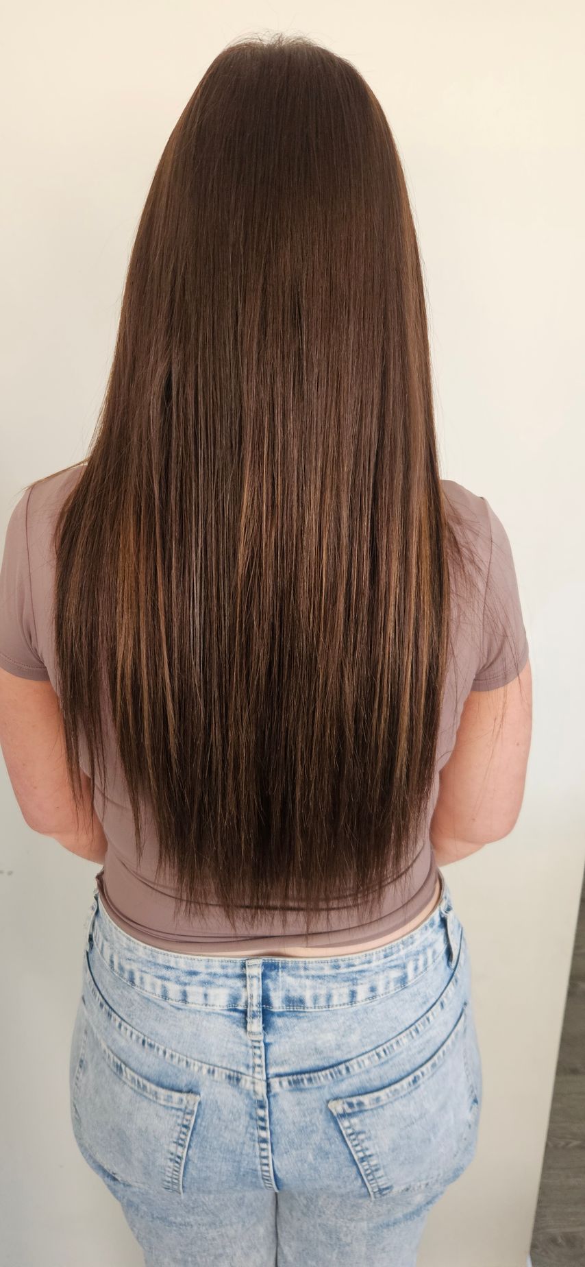 Woman with long, straight brown hair wearing a pink top and light-wash jeans, standing in front of a wall.