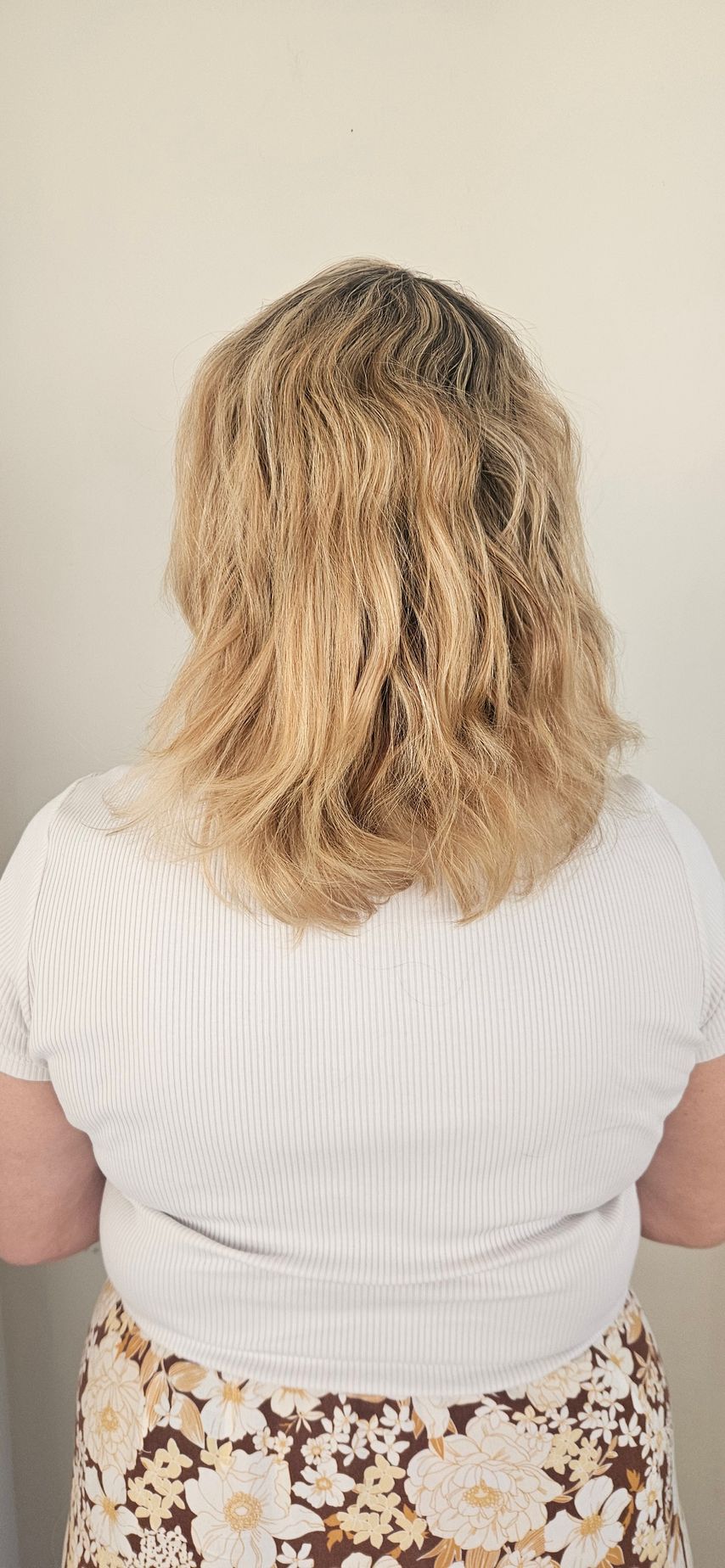 A person with blonde, curly hair wearing a white shirt and floral skirt, seen from the back.