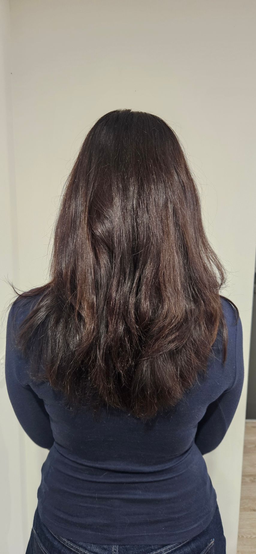 Person with dark, wavy hair from the back, wearing a blue shirt. White wall in the background.
