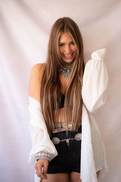 Woman with long brown hair smiles broadly at the camera in a well-lit room.