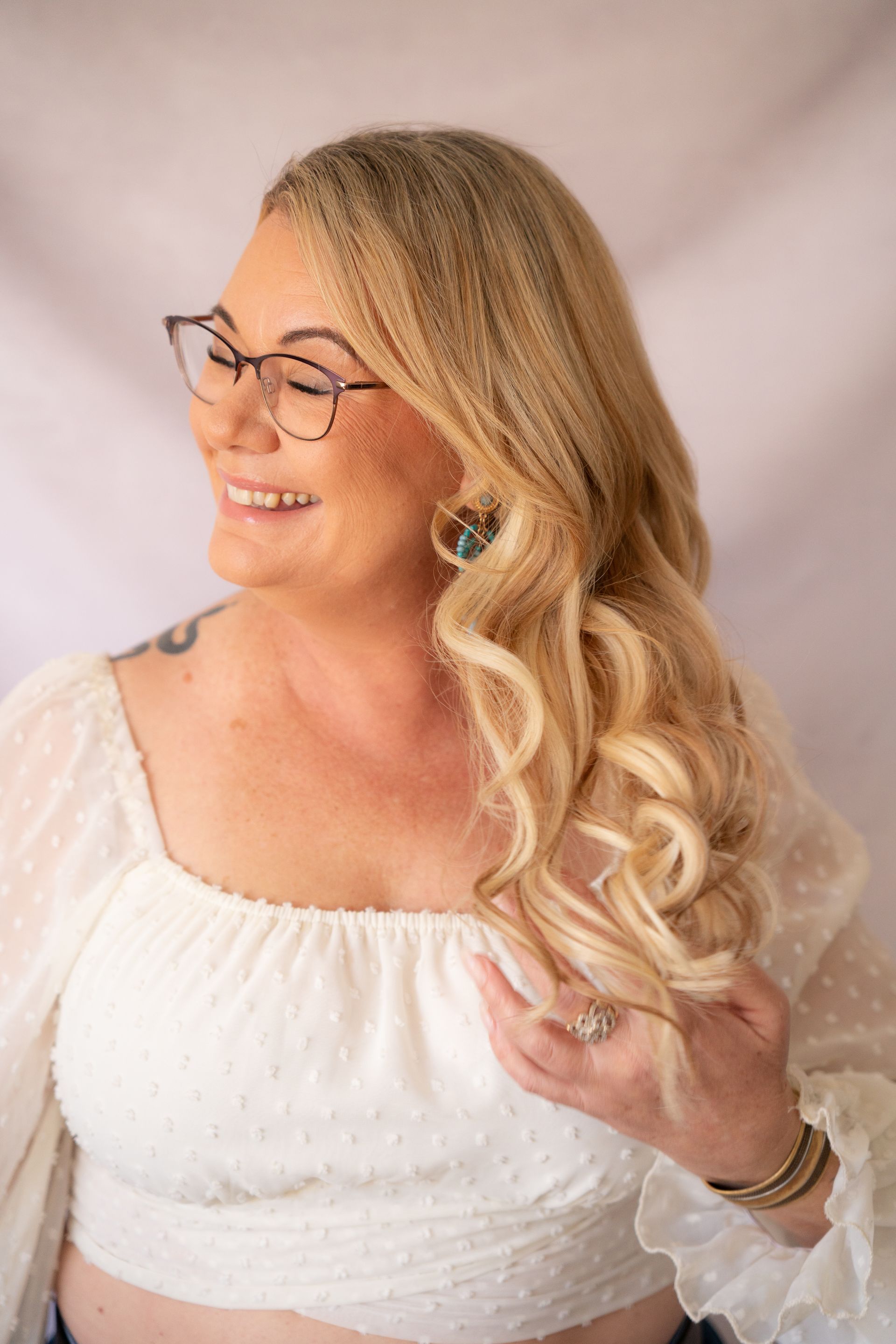 Blonde woman smiling, wearing glasses and white top, hair in curls, jewelry visible.
