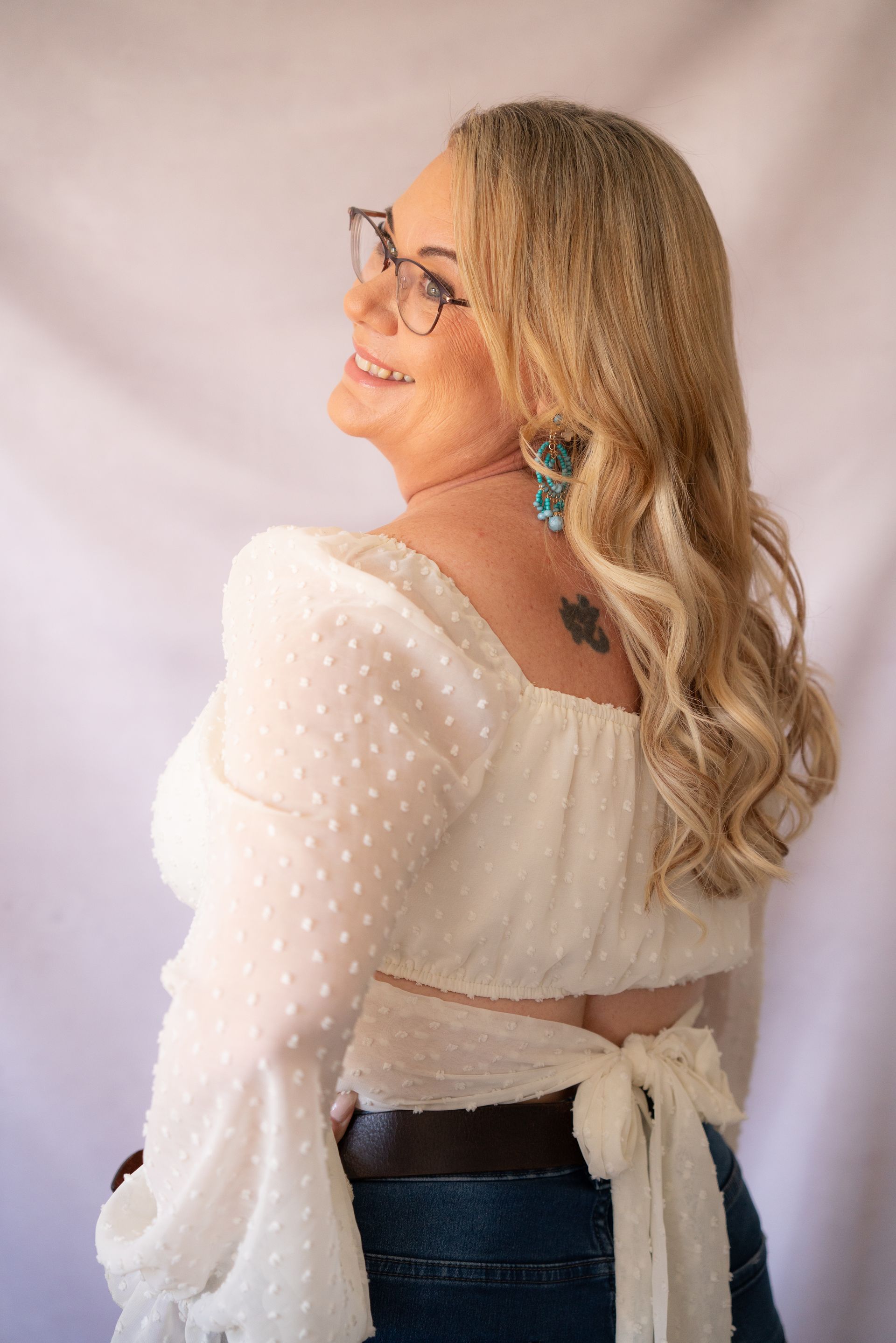 Woman with blonde hair, wearing glasses, smiling, in a white top and jeans, looking upward.