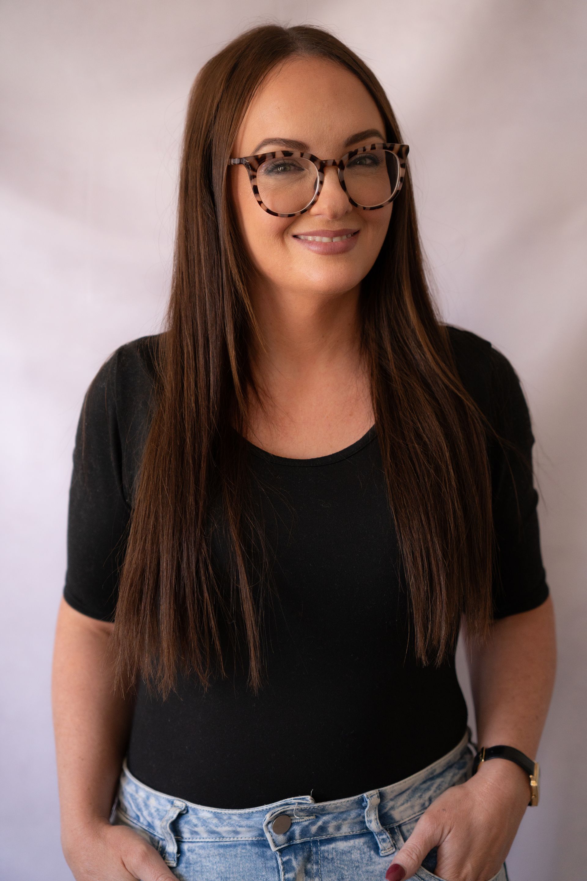 Woman with long brown hair, wearing glasses, black shirt, and jeans, smiling.