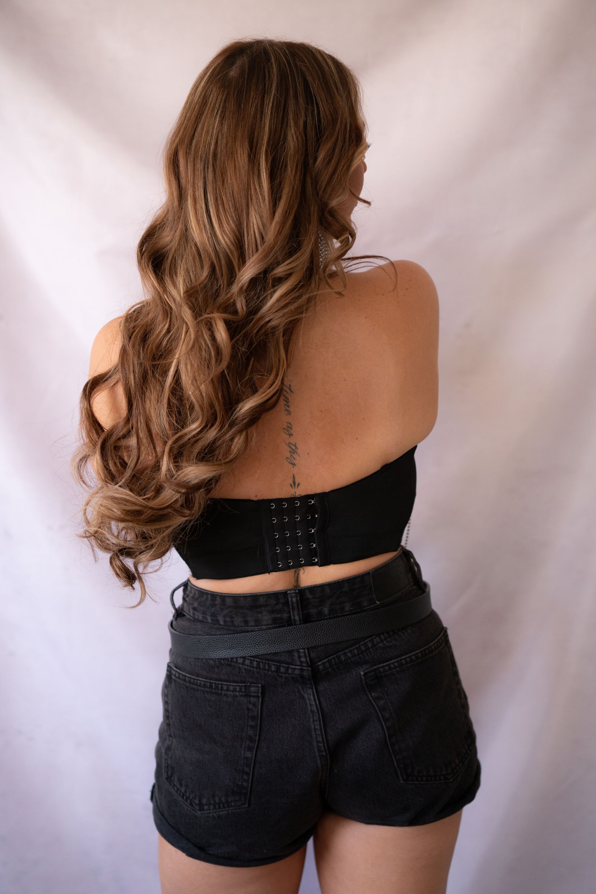 Woman from behind, wearing black crop top and shorts, long wavy brown hair, against a white backdrop.
