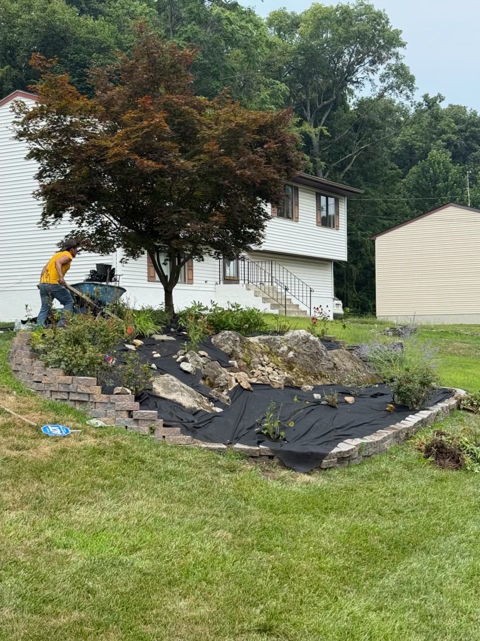 Man using a wheelbarrow to work in a garden bed in front of a white house.