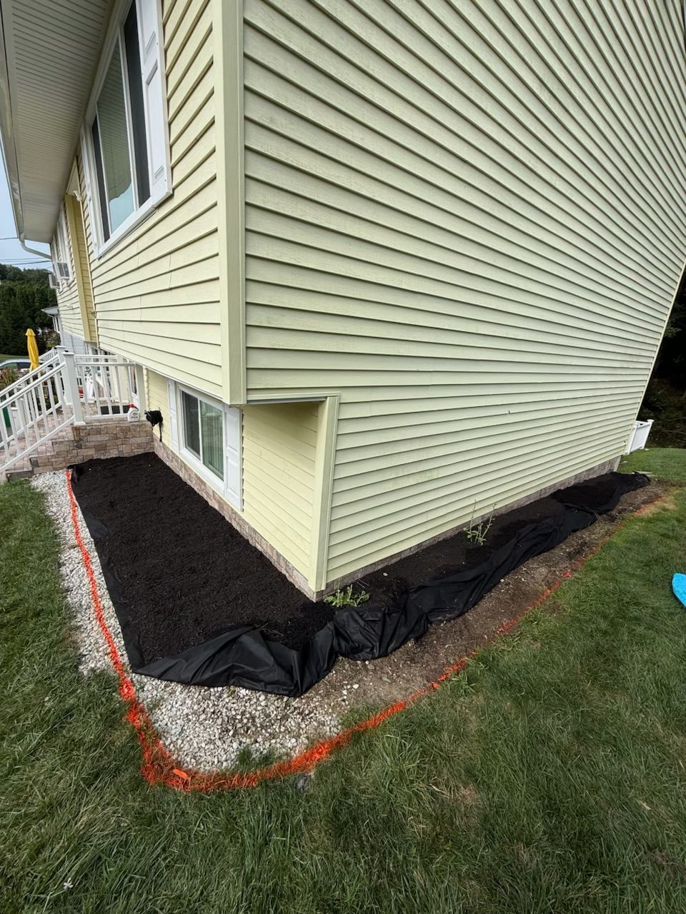 House with a mulch-filled bed outlined in orange, along the foundation. Black fabric lines the bed; green grass surrounds.