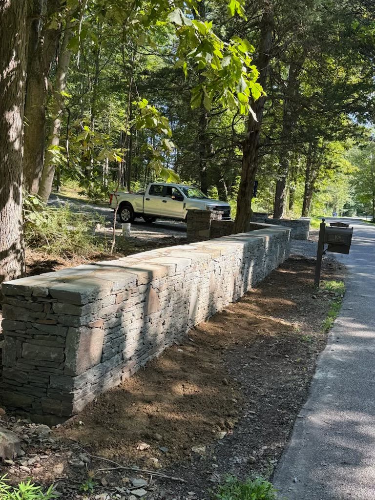 Stone retaining wall with dirt bed next to a road, trees, and a parked pickup truck.
